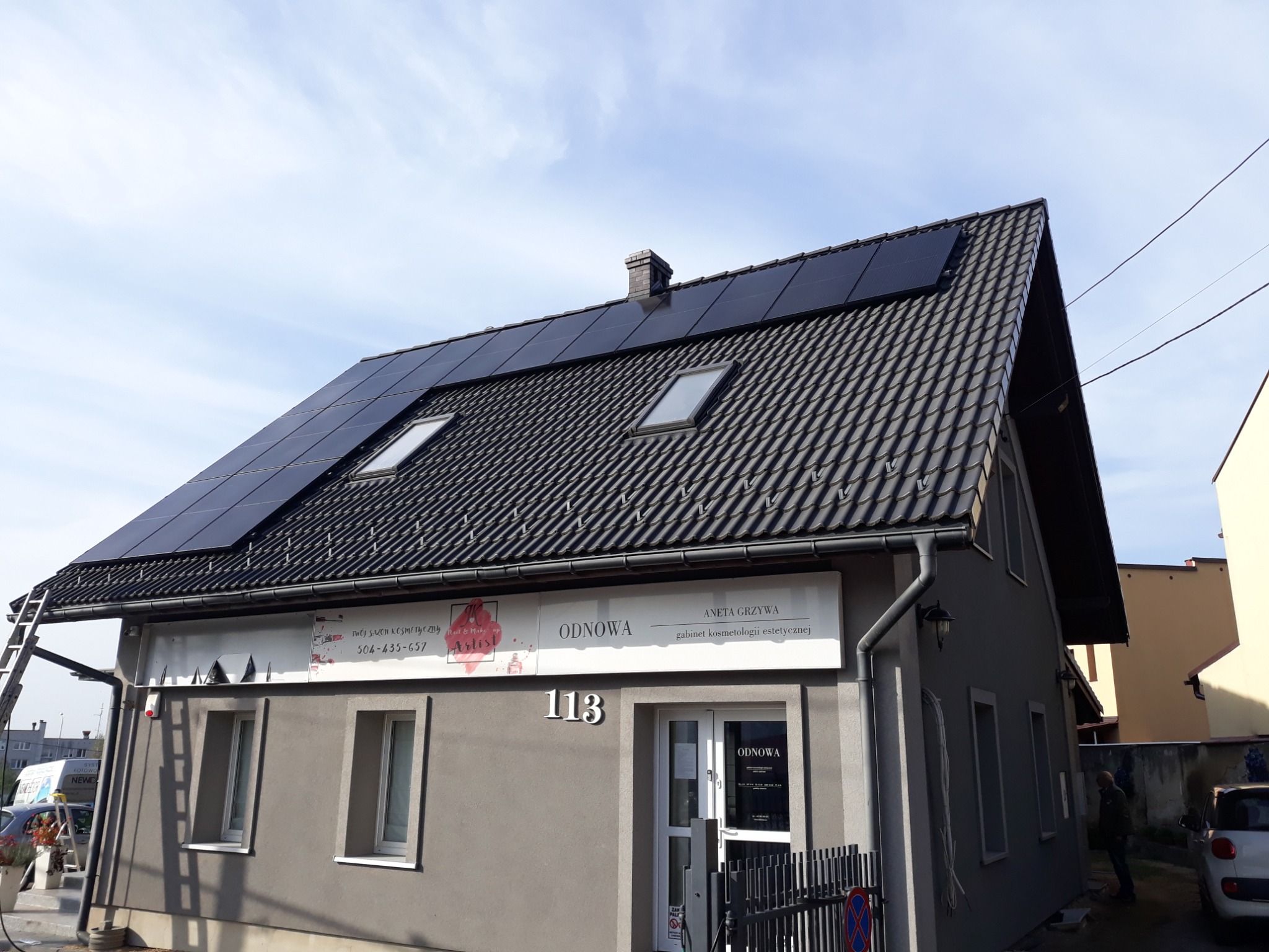 Dom z panelami słonecznymi na dachu, widoczne okna dachowe i drabina oparta o budynek, na którym znajduje się szyld gabinetu kosmetycznego.