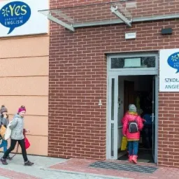 Wejście do szkoły językowej 'Yes school of English' z uczniami wchodzącymi do środka, widoczny szyld z nazwą szkoły w języku angielskim i polskim na ceglanej ścianie budynku.