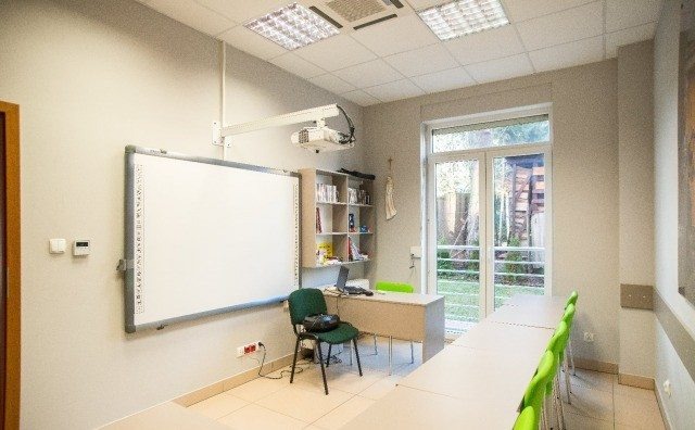 Jasna sala lekcyjna z interaktywną tablicą, projektorem na uchwycie, biurkiem, krzesłem i rzędem stołów z zielonymi krzesłami, widok na ogród przez przeszklone drzwi.