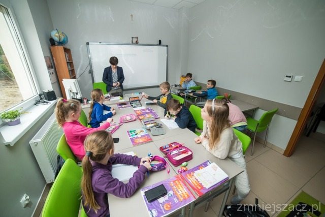 Sala lekcyjna z grupą dzieci siedzących przy stołach i kobietą stojącą przy tablicy interaktywnej, widok z perspektywy ucznia.