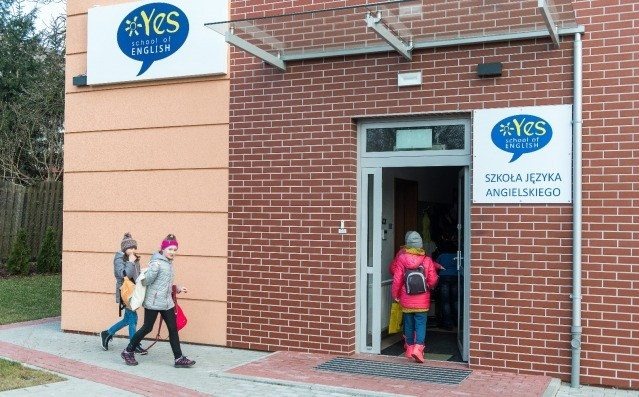 Wejście do szkoły językowej 'Yes school of English' z uczniami wchodzącymi do środka, widoczny szyld z nazwą szkoły w języku angielskim i polskim na ceglanej ścianie budynku.