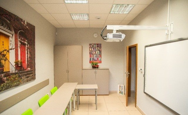Wnętrze sali lekcyjnej we Wrocławiu z rzutnikiem przymocowanym do sufitu, tablicą interaktywną, stołami, zielonymi krzesłami i dekoracją ścienną przedstawiającą kolorowe drzwi.