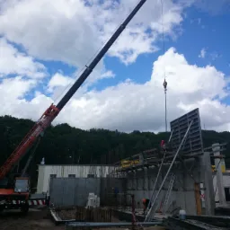 Żuraw wieżowy w trakcie podnoszenia elementu szalunku na placu budowy, widoczni pracownicy w kaskach, w tle las i błękitne niebo z chmurami.