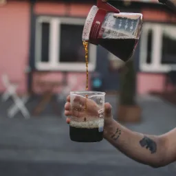 Ręka z tatuażami nalewa kawę z karafki do szklanego naczynia laboratoryjnego na tle budynku.