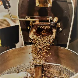 Proces prażenia ziaren kawy w przemysłowym piecu z widocznym termometrem i ręką obsługującą mechanizm. Ziarna wsypywane do metalowego bębna z mieszadłem.