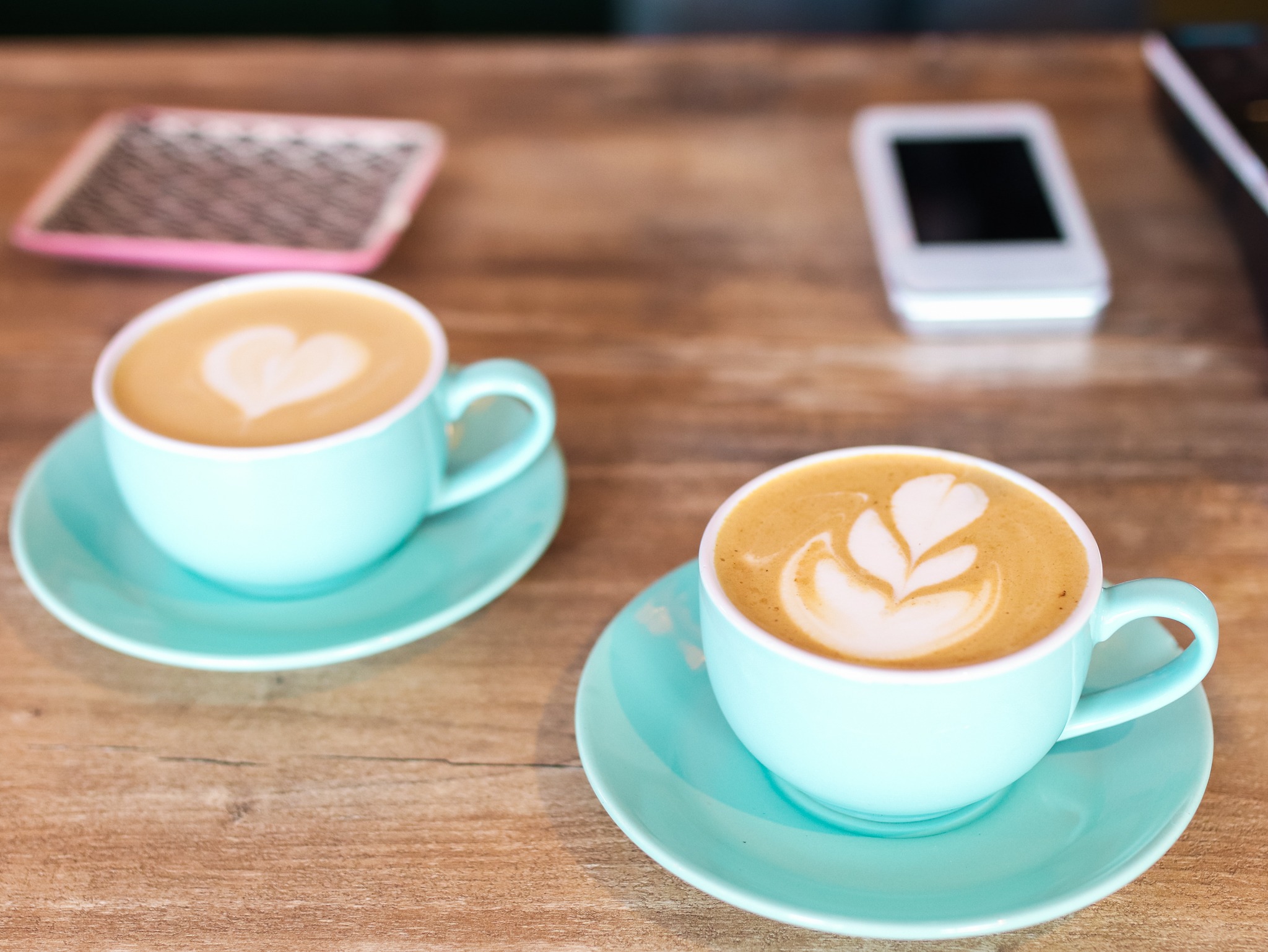 Dwie turkusowe filiżanki cappuccino z latte art na drewnianym stole, obok etui na telefon i terminal płatniczy.