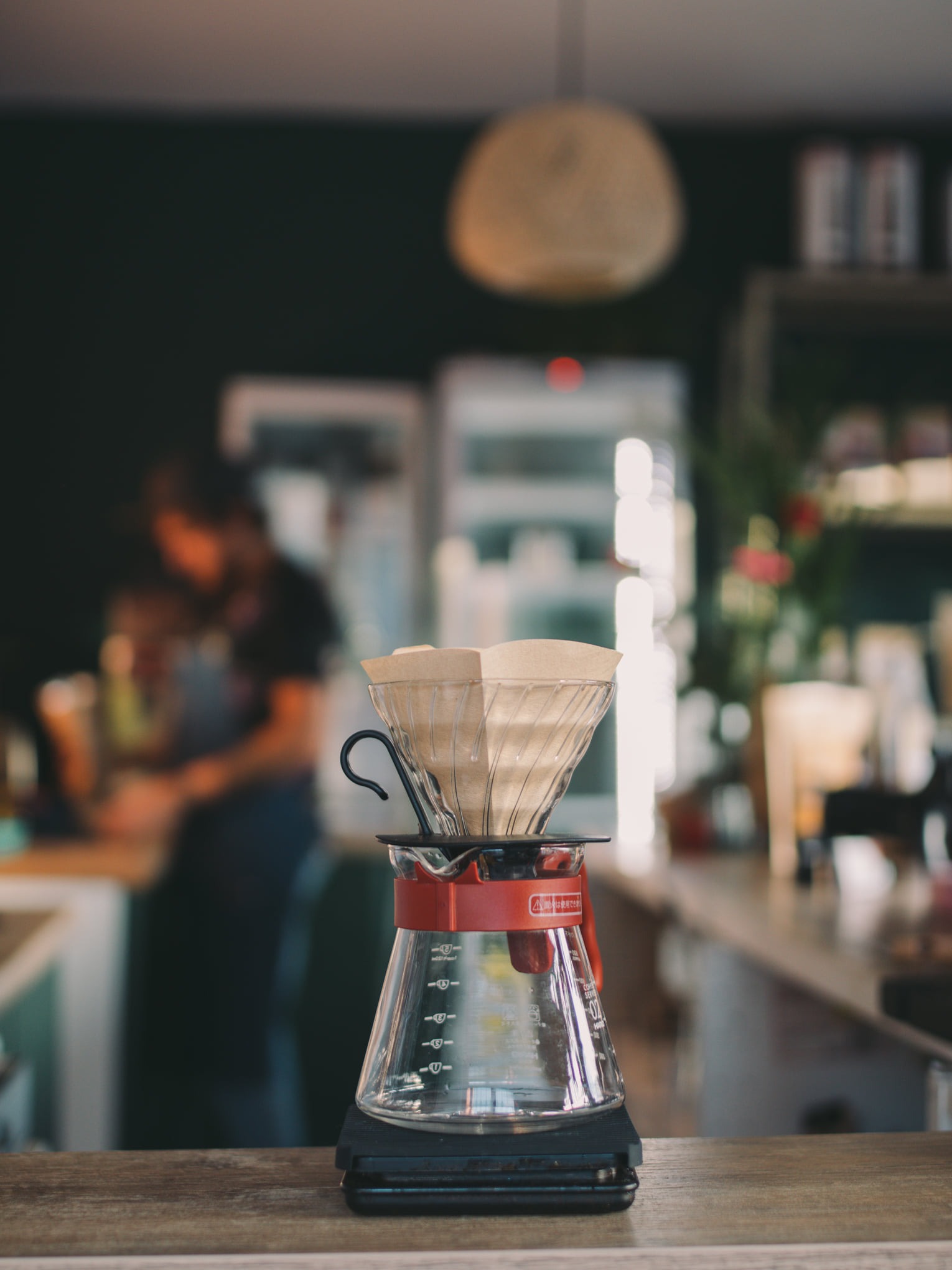 Szklany zaparzacz do kawy typu drip z papierowym filtrem, umieszczony na czarnej wadze elektronicznej, w tle widoczna kawiarnia z pracującymi osobami.