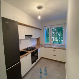 Nowoczesna kuchnia w bieli i czerni z drewnopodobnym blatem, górne szafki bez uchwytów, dolne z czarnymi krawędziowymi, na podłodze dwie butelki wody