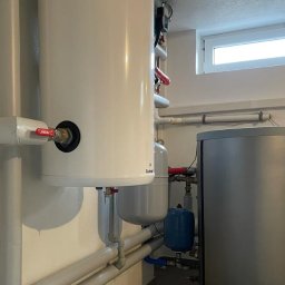 Auto-Maro Sp. z o.o. - Biały zasobnik ciepłej wody użytkowej z zaworem Ferro, szare rury instalacyjne, niebieski zbiornik przeponowy i szary zbiornik akumulacyjny w kotłowni z małym oknem.