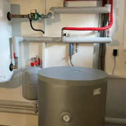 Instalacja kotłowni z dwoma zbiornikami, rurami w izolacji termicznej i panelem sterowania, widoczne manometry i zawory.