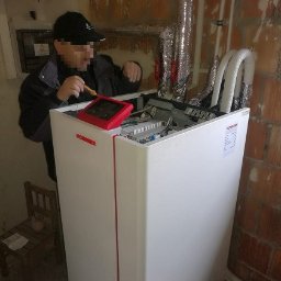 Auto-Maro Sp. z o.o. - Instalator w czapce diagnozuje pompę ciepła Ochsner za pomocą miernika, widoczne rury, ceglana ściana i skrzynka elektryczna w tle.