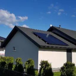 Panele fotowoltaiczne zamontowane na dachu domu jednorodzinnego z ciemną dachówką, widok z poziomu gruntu w słoneczny dzień.