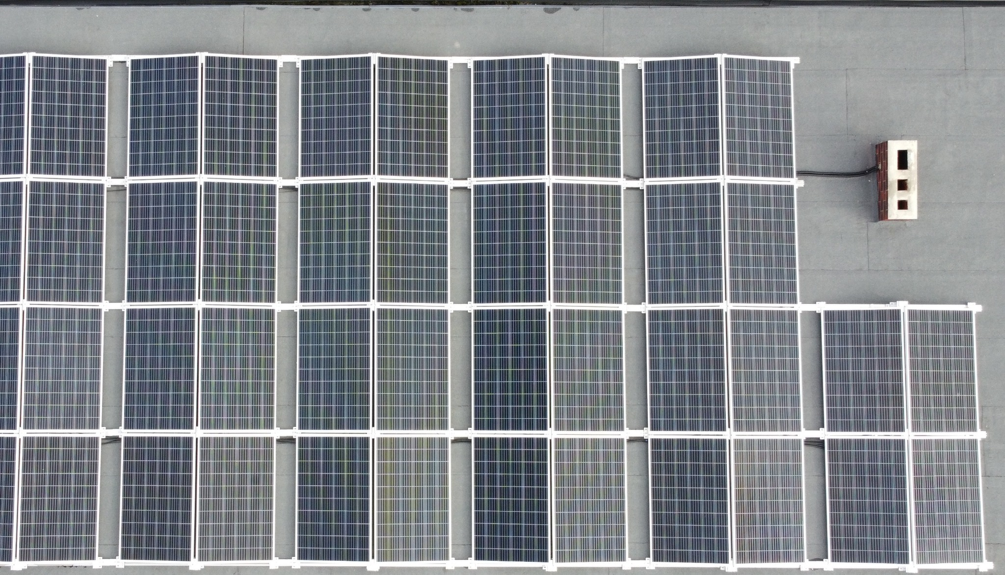 Widok z góry na panele słoneczne z białymi ramami, ułożone rzędami na szarym dachu, z widoczną instalacją elektryczną przy kominie wentylacyjnym.