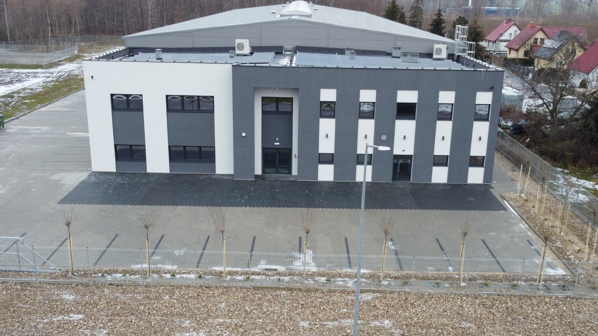 Nowoczesny budynek biurowy z panelami słonecznymi na dachu i klimatyzatorami, widok z góry w pochmurny dzień, z szarym brukowanym podjazdem i młodymi drzewkami wzdłuż ogrodzenia.