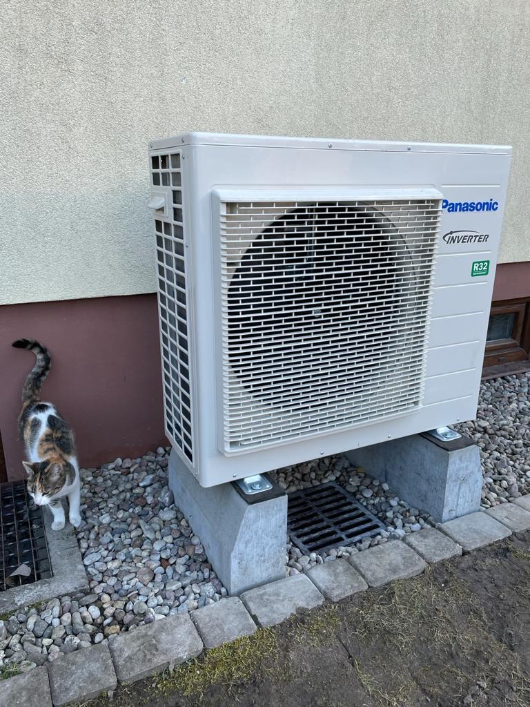Zewnętrzna jednostka pompy ciepła Panasonic Inverter R32 na betonowych podstawach, z kotem przy kratce ściekowej i kamiennym podłożu.