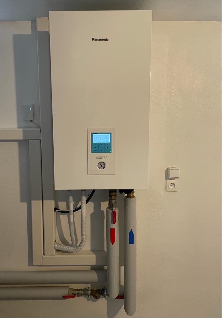 Biała pompa ciepła Panasonic Aquarea zamontowana na ścianie, z widocznymi rurami ocieplonymi z oznaczeniami czerwonej i niebieskiej strzałki, oraz panel sterowania z wyświetlaczem LCD.