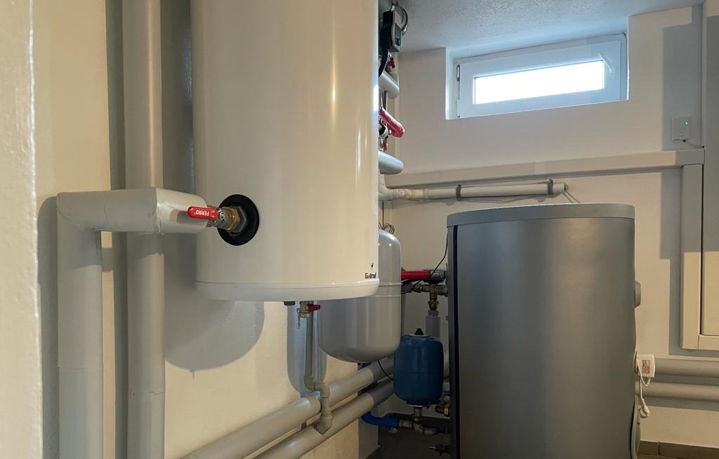 Biały zasobnik ciepłej wody użytkowej z zaworem Ferro, szare rury instalacyjne, niebieski zbiornik przeponowy i szary zbiornik akumulacyjny w kotłowni z małym oknem.