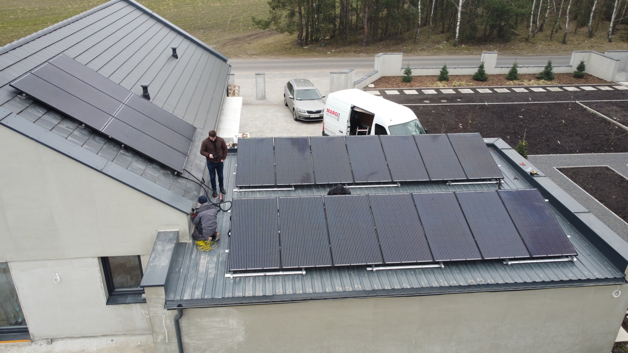 Montaż paneli słonecznych na dachu domu jednorodzinnego z widocznym samochodem i busem firmy instalacyjnej na posesji.