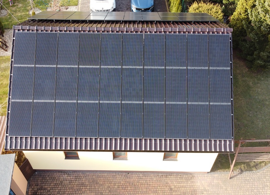 Widok z góry na dach domu z zamontowanymi panelami słonecznymi, dwa samochody zaparkowane obok, widoczny fragment ogrodu i kostki brukowej.