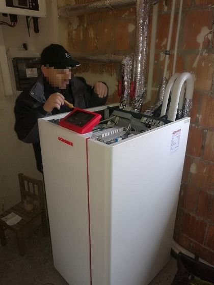 Instalator w czapce diagnozuje pompę ciepła Ochsner za pomocą miernika, widoczne rury, ceglana ściana i skrzynka elektryczna w tle.