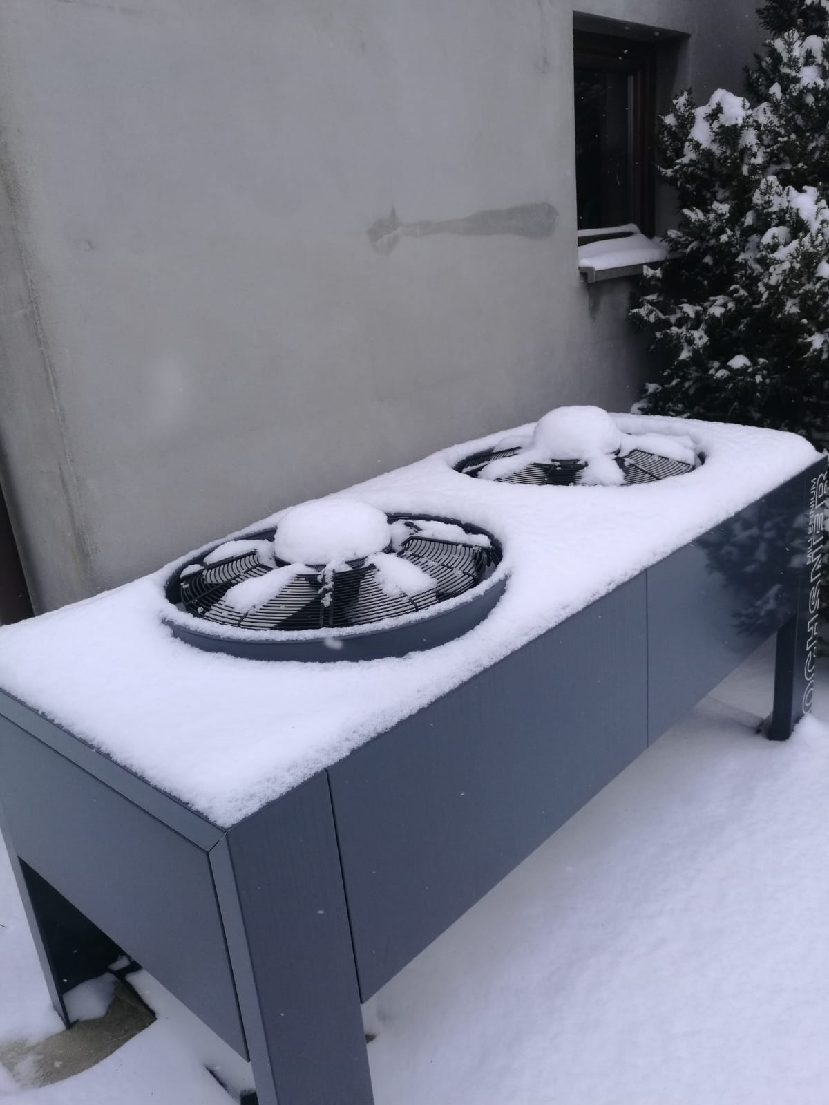 Szara pompa ciepła z dwoma wentylatorami, częściowo zasypana śniegiem, stoi na zewnątrz budynku w Koninie, w tle widoczna ściana i krzew.