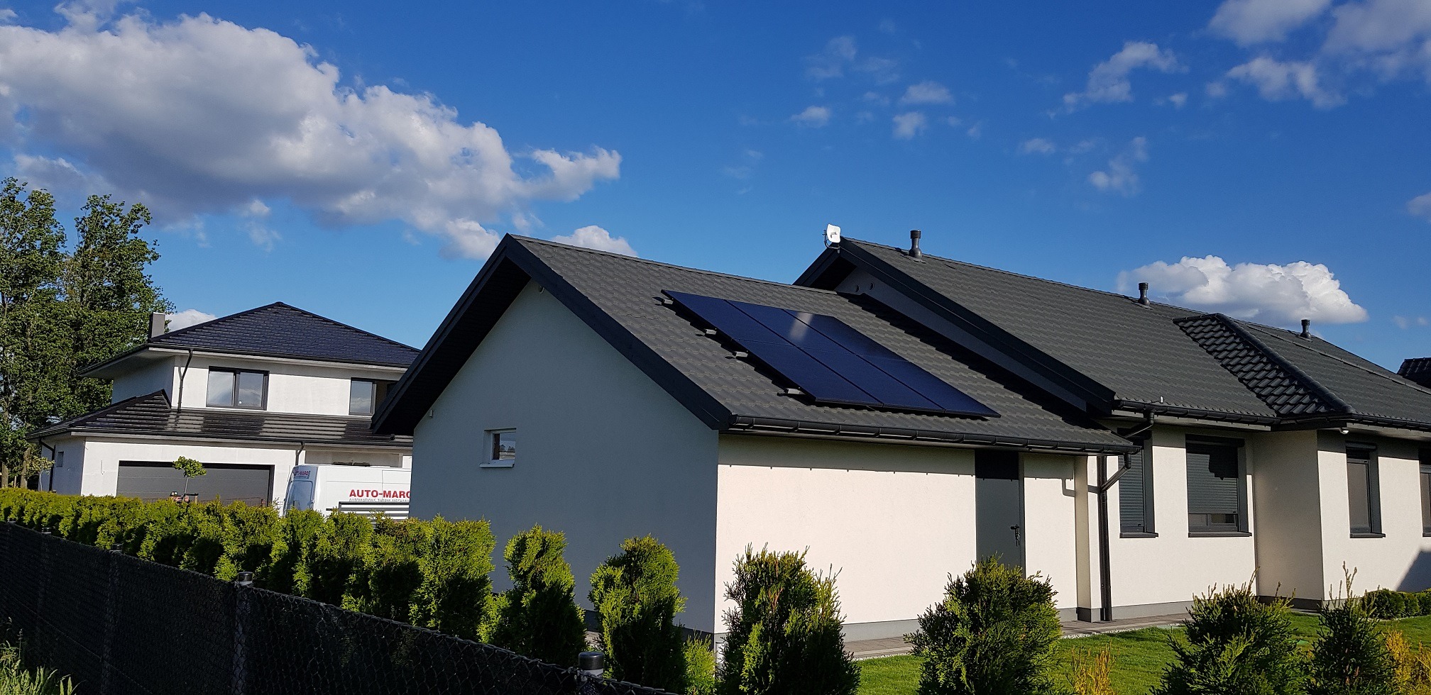 Panele fotowoltaiczne zamontowane na dachu domu jednorodzinnego z ciemną dachówką, widok z poziomu gruntu w słoneczny dzień.