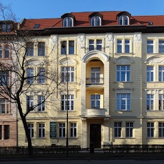 Fasada kamienicy w stylu secesyjnym z żółtą elewacją, ozdobnymi balkonami i czerwonym dachem, widoczna z poziomu ulicy w słoneczny dzień.