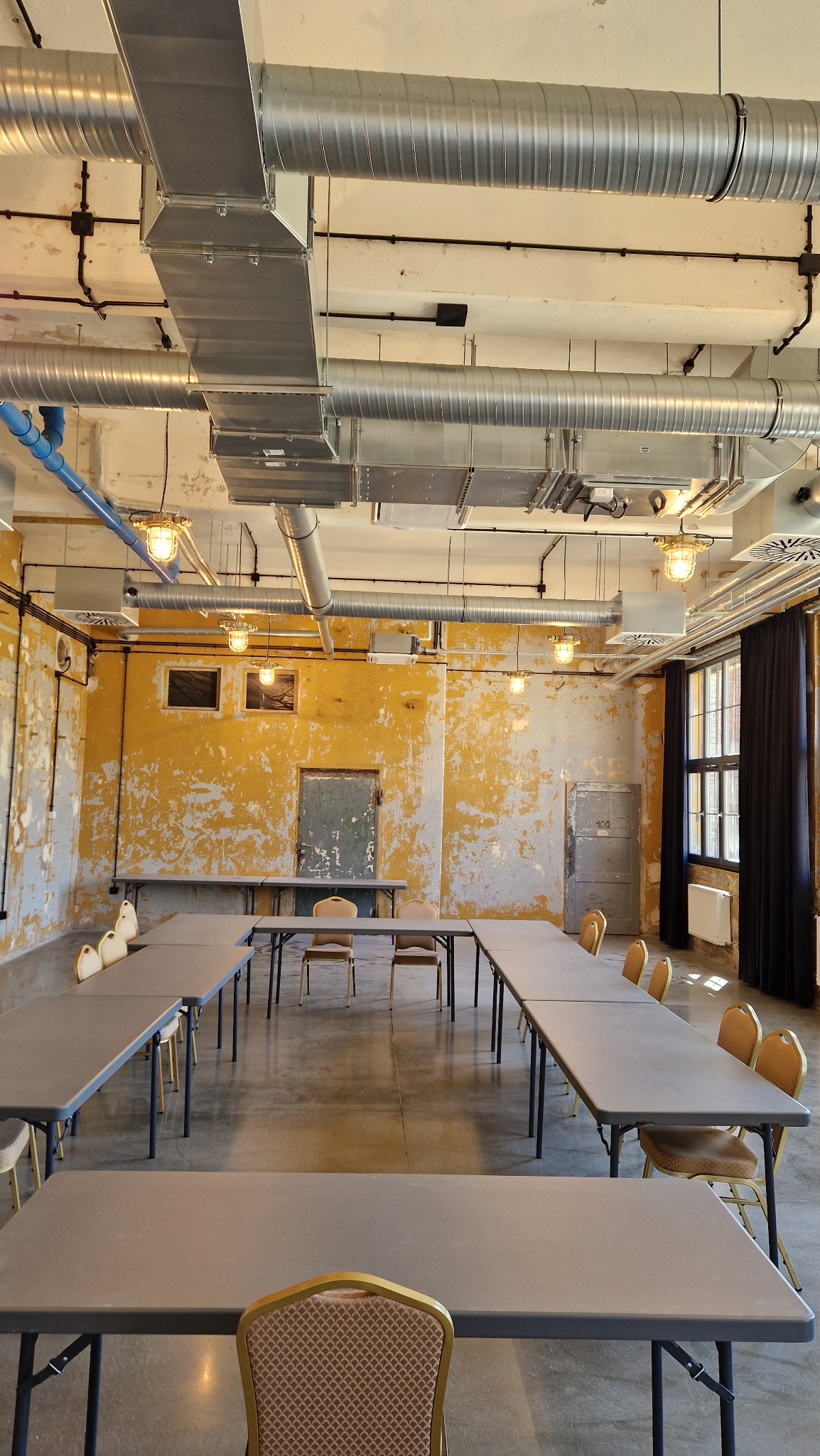 Sala konferencyjna w stylu industrialnym z odsłoniętą instalacją wentylacyjną pod sufitem, żółtymi ścianami z odpryskującą farbą i długimi stołami ustawionymi w kształt litery U.