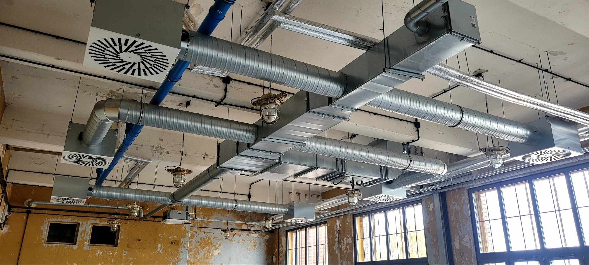 System kanałów wentylacyjnych i klimatyzacyjnych z okrągłymi i prostokątnymi rurami pod sufitem w industrialnym wnętrzu z oknami i lampami w metalowych koszach.