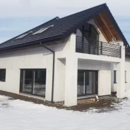 Nowoczesny dom murowany z płaskim dachem i balkonem, wykończony białą elewacją, widoczny w zimowym krajobrazie ze śniegiem, z czarnymi ramami okien i balustradą balkonu.