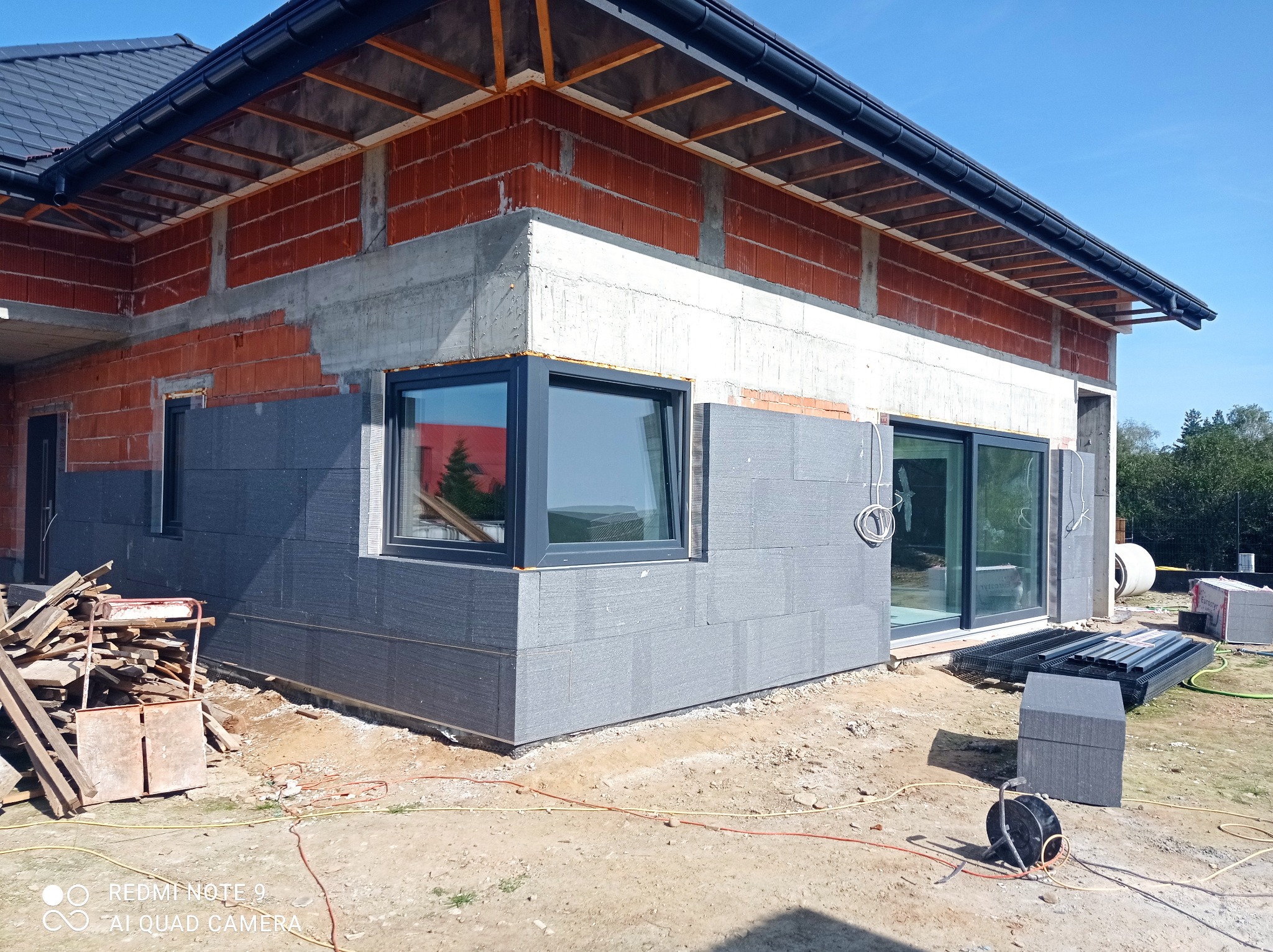 Surowy stan budynku jednorodzinnego z czerwonej cegły, z zamontowanymi oknami i częściowo wykonaną izolacją zewnętrzną z płyt grafitowych, widoczne materiały budowlane i prowizoryczne składowisko...