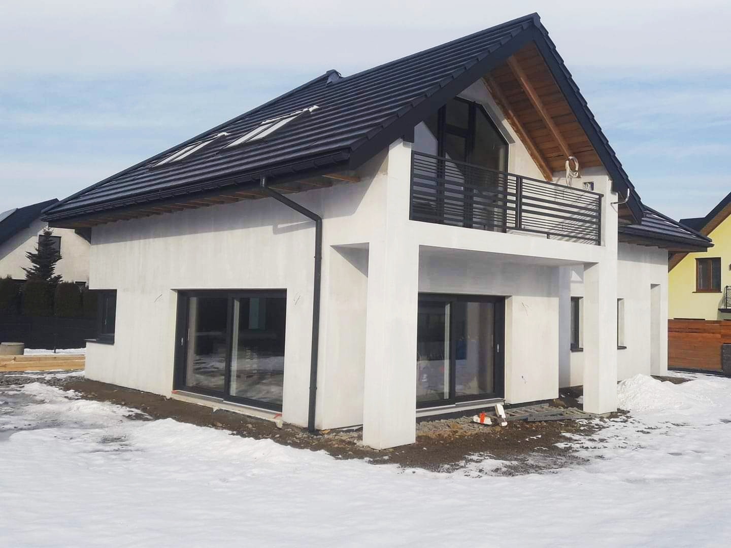 Nowoczesny dom murowany z płaskim dachem i balkonem, wykończony białą elewacją, widoczny w zimowym krajobrazie ze śniegiem, z czarnymi ramami okien i balustradą balkonu.