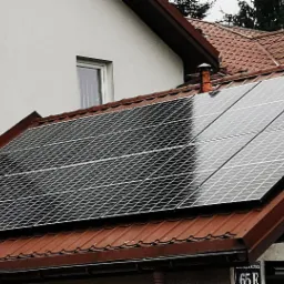 Panele słoneczne na dachu domu z brązową dachówką i oknem z kratą, widoczny klimatyzator na ścianie budynku.