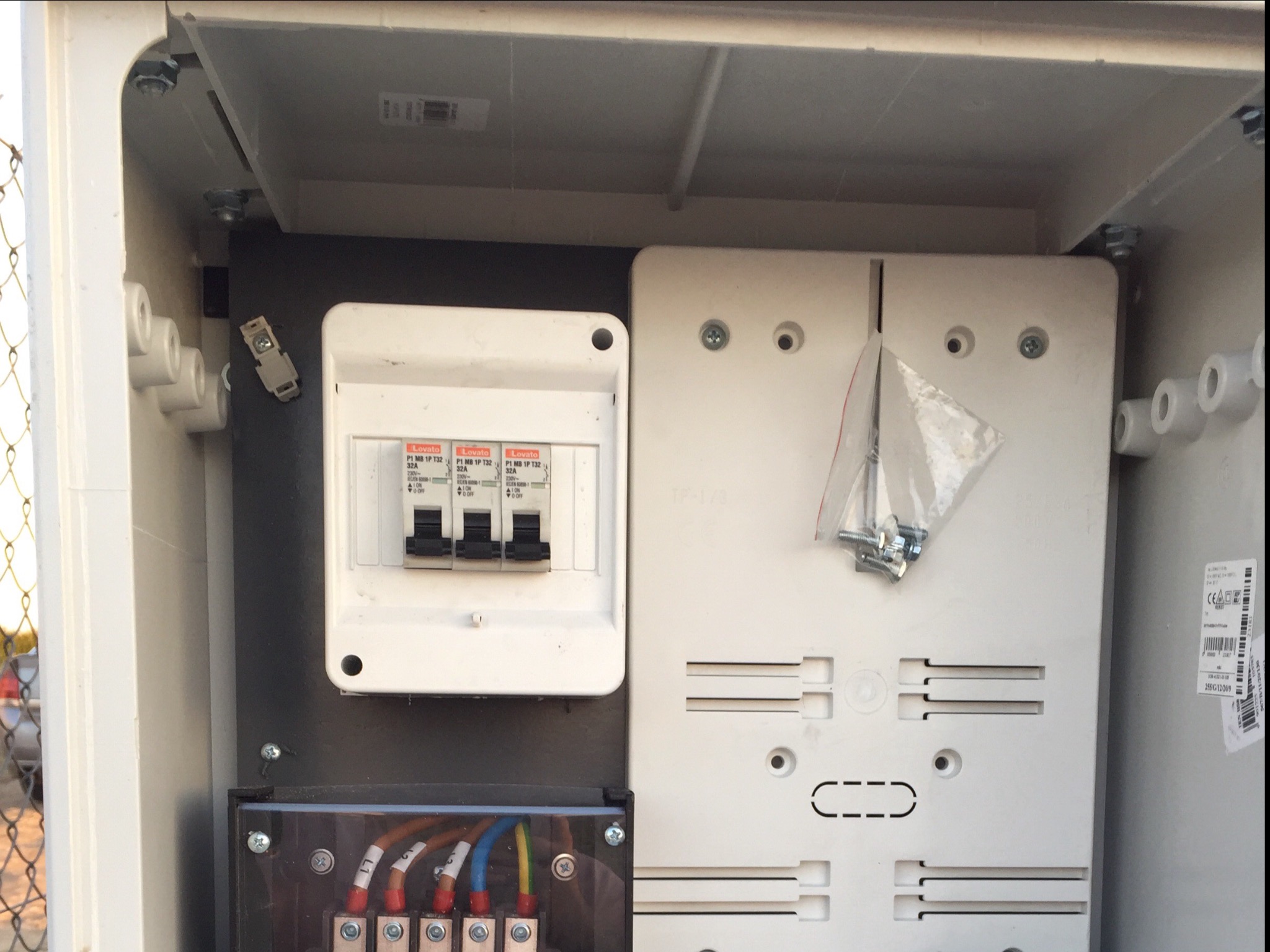 Wnętrze skrzynki elektrycznej z zamontowanymi bezpiecznikami Lovato 32A, przewodami oznaczonymi kolorami, oraz woreczkiem ze śrubami montażowymi, widoczne elementy instalacji elektrycznej.