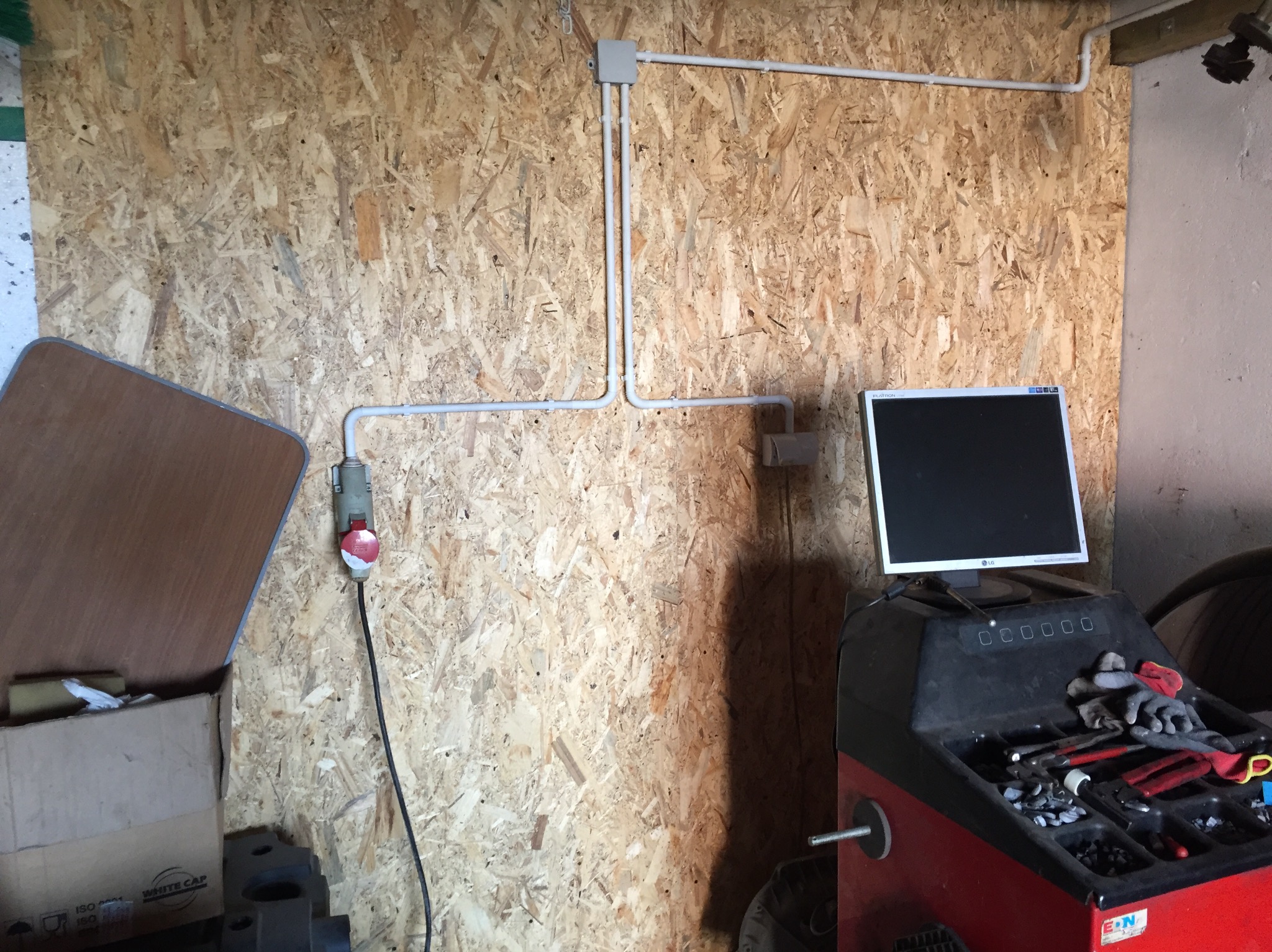 Szara instalacja elektryczna natynkowa z puszkami i gniazdem siłowym na ścianie z płyty OSB, obok stanowisko komputerowe do wyważania kół z narzędziami i rękawicami roboczymi.