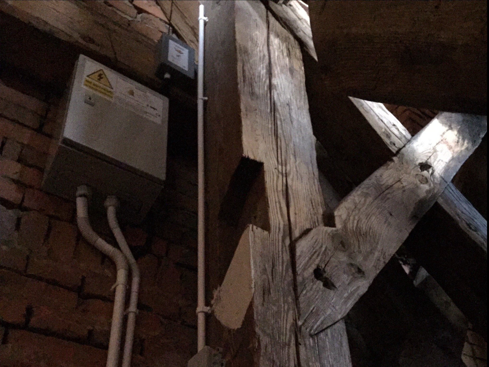 Szara skrzynka elektryczna z symbolem ostrzegawczym na ceglanej ścianie poddasza, z widocznymi rurami i drewnianą konstrukcją dachu.