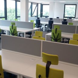 Przedsiębiorstwo wielobranżowe Viggo - Nowoczesne biuro z rzędami białych biurek oddzielonych szarymi przegrodami i ergonomicznymi krzesłami w odcieniach zieleni i żółci, widok z perspektywy pracownika.