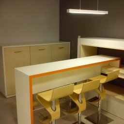 Przedsiębiorstwo wielobranżowe Viggo - Nowoczesny barek z białym blatem i pomarańczowym akcentem, uzupełniony trzema hokerami o organicznych kształtach, z szafką w tle i podłużną lampą sufitową.