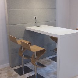 Przedsiębiorstwo wielobranżowe Viggo - Biały blat stołu z wbudowanym gniazdem elektrycznym, umieszczony przy szarej, tapicerowanej ścianie w biurze w Krakowie, z dwoma nowoczesnymi krzesłami barowymi z giętego drewna.