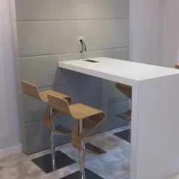 Biały blat stołu z wbudowanym gniazdem elektrycznym, umieszczony przy szarej, tapicerowanej ścianie w biurze w Krakowie, z dwoma nowoczesnymi krzesłami barowymi z giętego drewna.