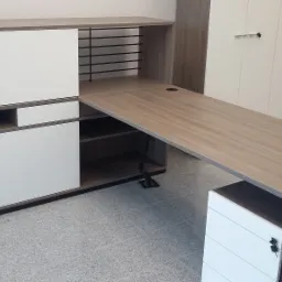 Biurko z szafkami w kolorze drewna i bieli, z widocznymi półkami i otworami na kable, na tle białych szaf.