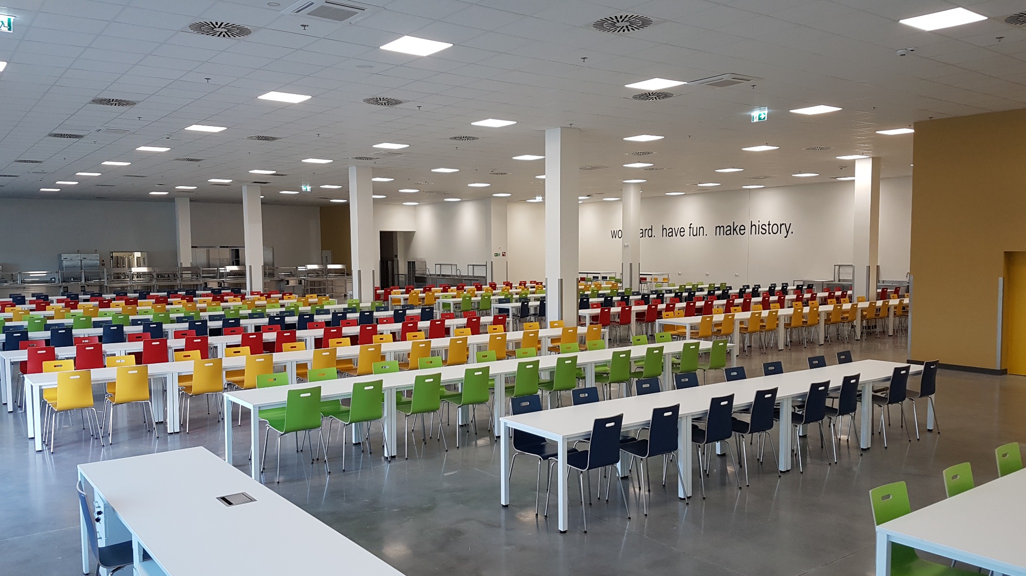 Przestronna sala jadalna z rzędami białych stołów i krzeseł w kolorach czerwonym, żółtym, zielonym i granatowym; na ścianie napis 'Work hard. Have fun. Make history.'