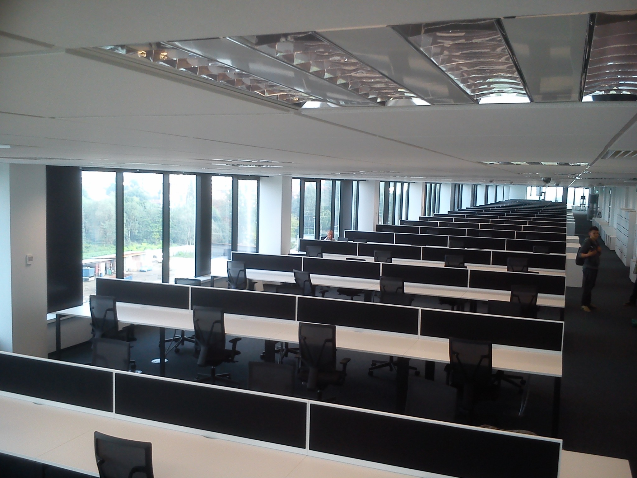 Długa perspektywa rzędu biurek z czarnymi krzesłami i panelami działowymi w przestronnym biurze z oknami od podłogi do sufitu.