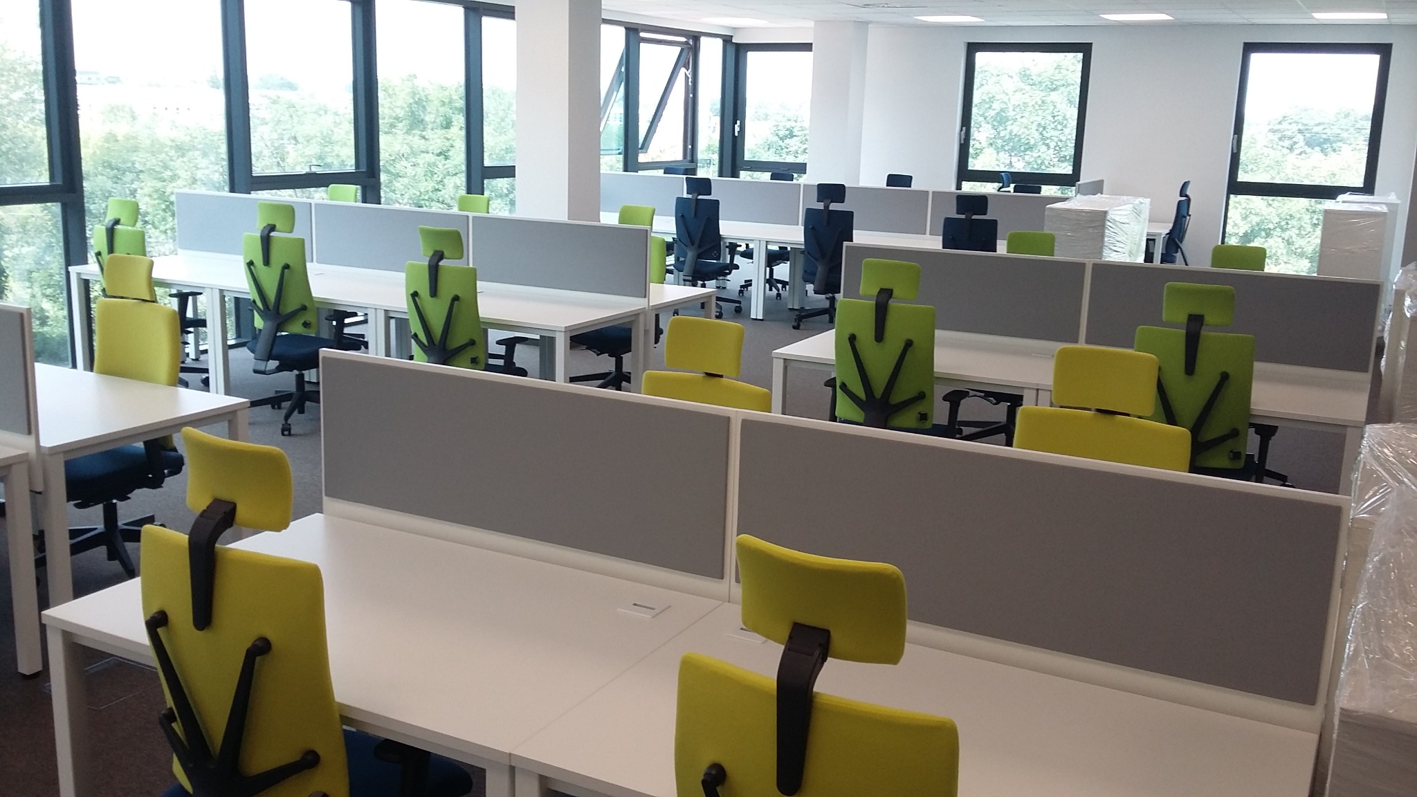 Nowoczesne biuro z rzędami białych biurek oddzielonych szarymi przegrodami i ergonomicznymi krzesłami w odcieniach zieleni i żółci, widok z perspektywy pracownika.