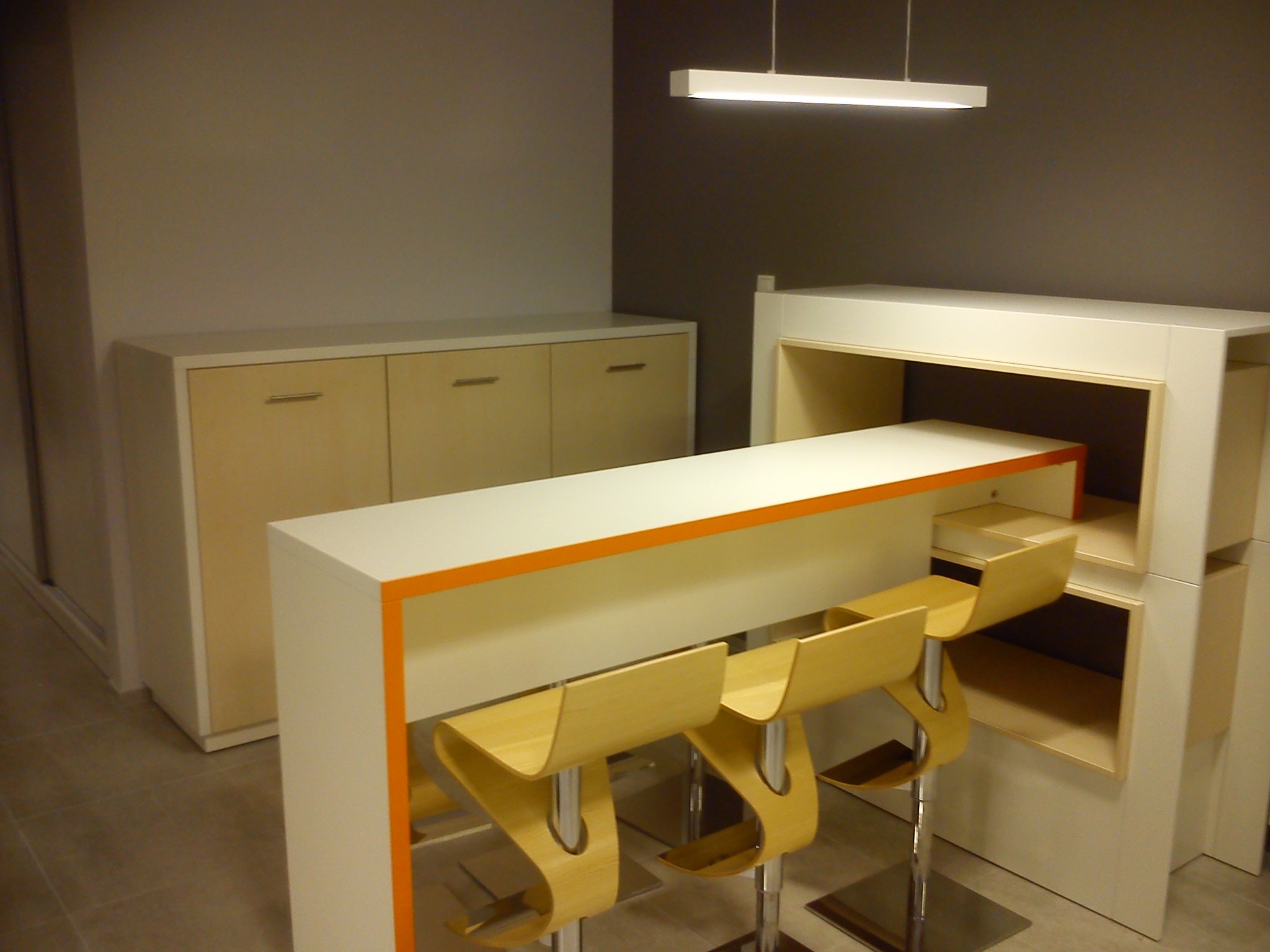 Nowoczesny barek z białym blatem i pomarańczowym akcentem, uzupełniony trzema hokerami o organicznych kształtach, z szafką w tle i podłużną lampą sufitową.