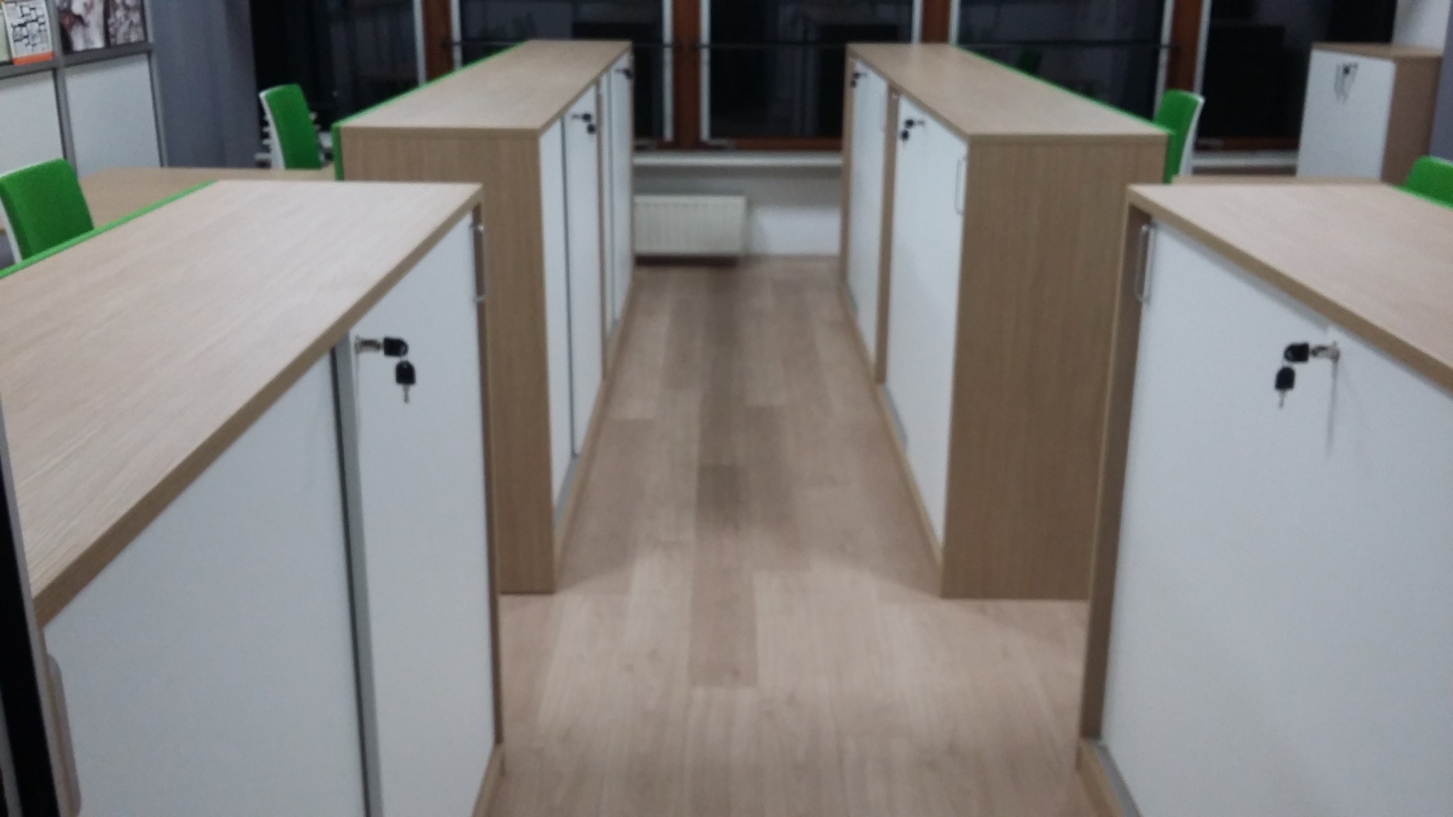 Długi korytarz biurowy z szeregiem szaf zamykanych na klucz, blat w kolorze jasnego drewna, białe fronty, widoczne krzesła biurowe w tle.