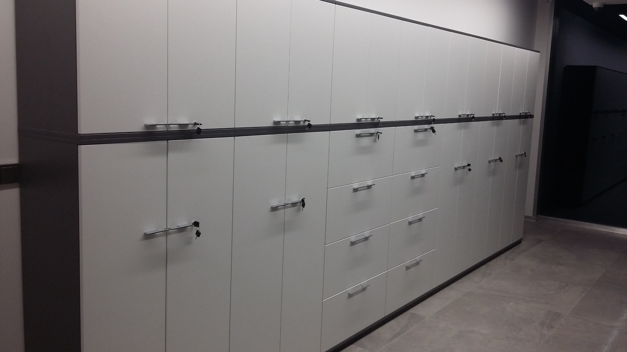 Długa ściana z białych szafek i szuflad z szarymi akcentami, zamkniętych na klucz, widok z perspektywy podłogi w biurze.