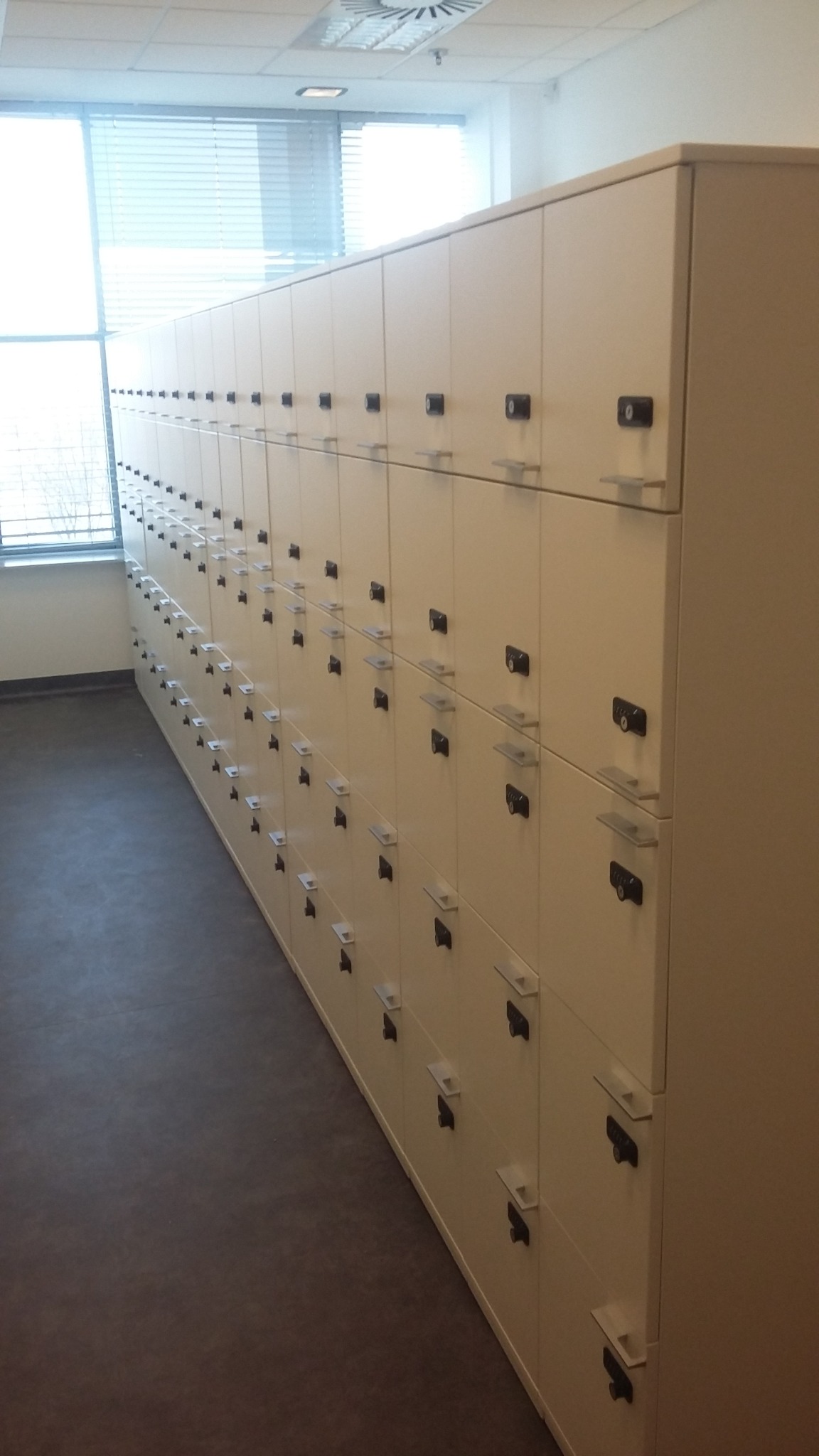 Rząd jasnych szafek z zamkami szyfrowymi w jasnym pomieszczeniu z oknem.