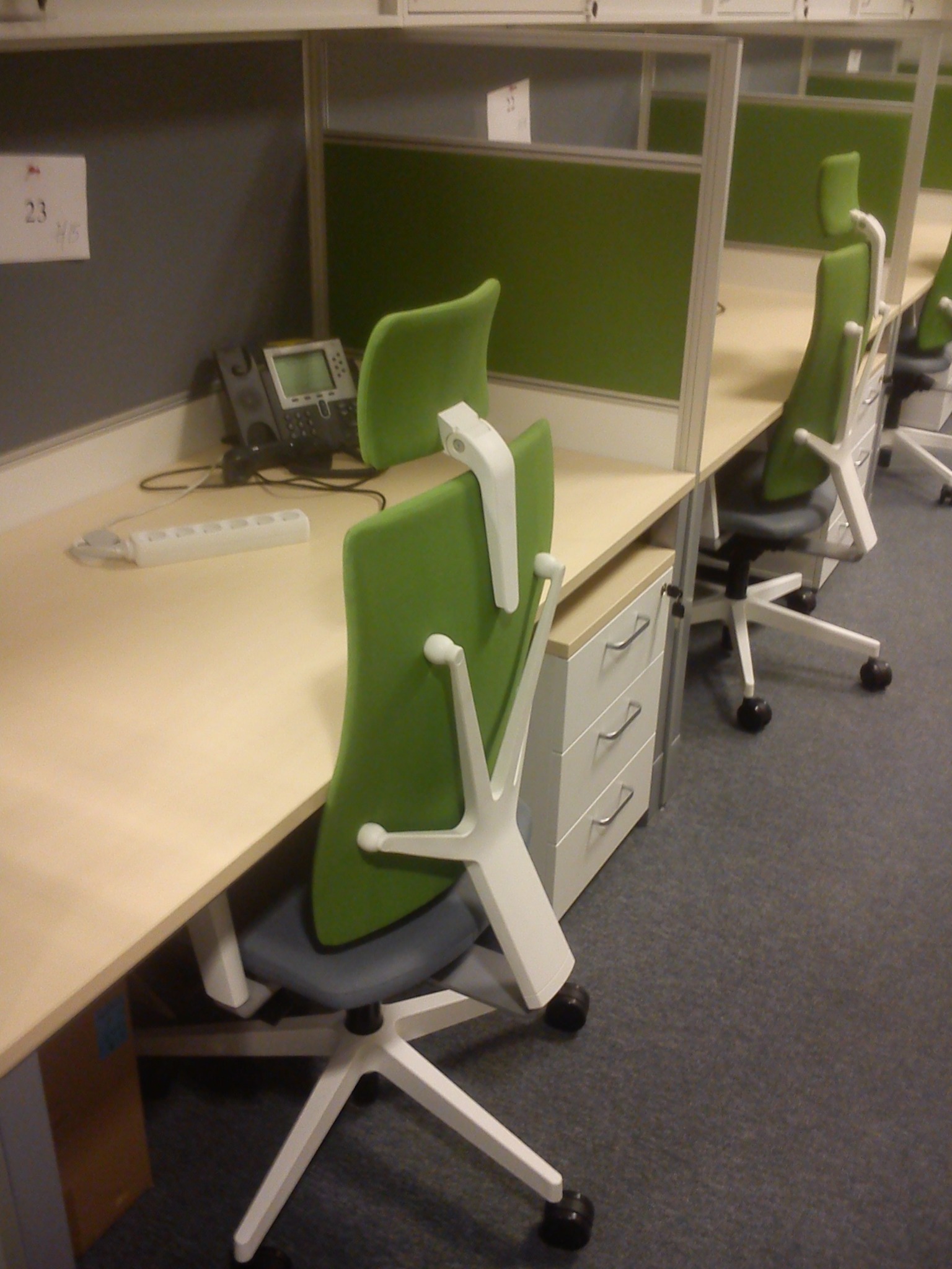 Rząd stanowisk pracy w biurze z biurkami z jasnego drewna, zielonymi ściankami działowymi i ergonomicznymi krzesłami z zielonym oparciem, widoczny telefon stacjonarny i przedłużacz na biurku.