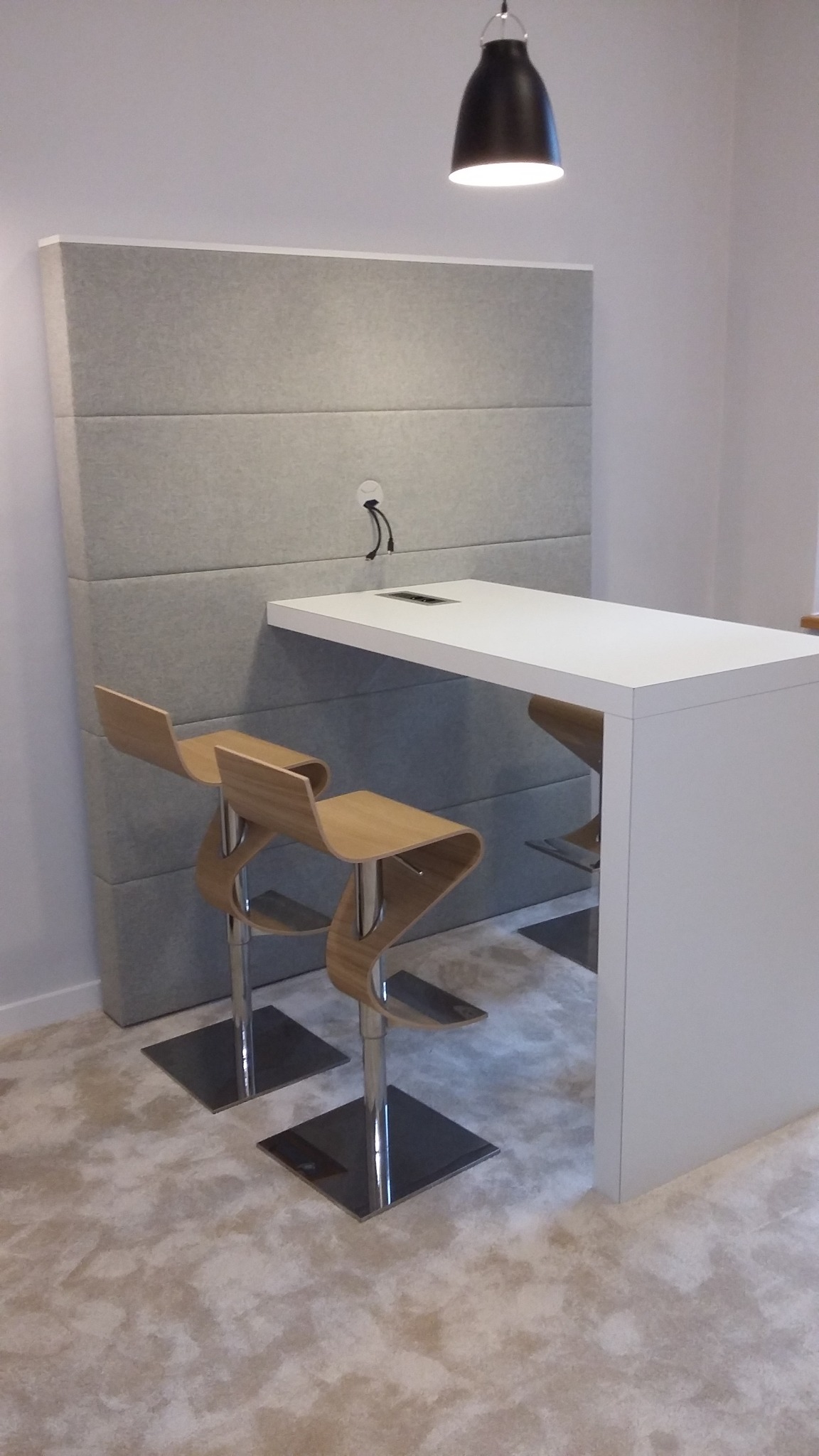 Biały blat stołu z wbudowanym gniazdem elektrycznym, umieszczony przy szarej, tapicerowanej ścianie w biurze w Krakowie, z dwoma nowoczesnymi krzesłami barowymi z giętego drewna.